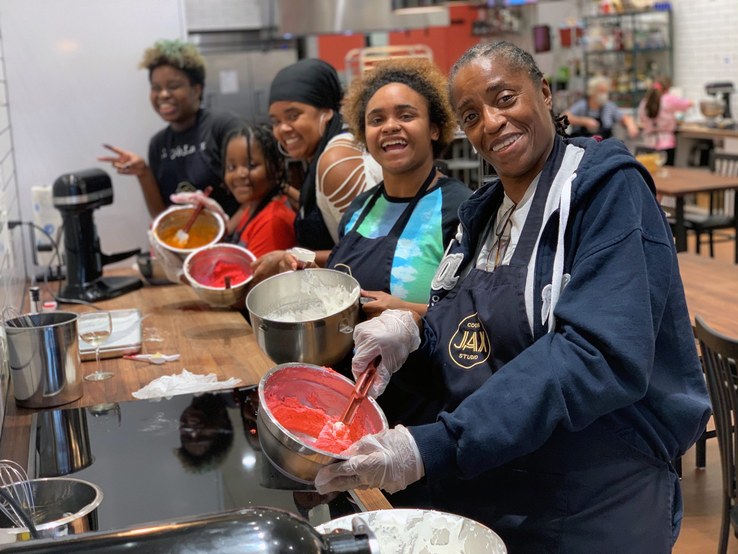 A family with three teenagers, one adult, and one child prepare red cake frosting in a cooking studio.