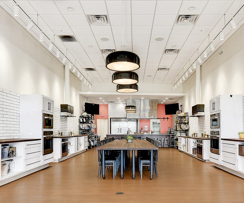 Interior of JAX Cooking Studio with a long, wooden communal table in the middle of the room and four cooking stations (with sinks and ovens) along the sides.