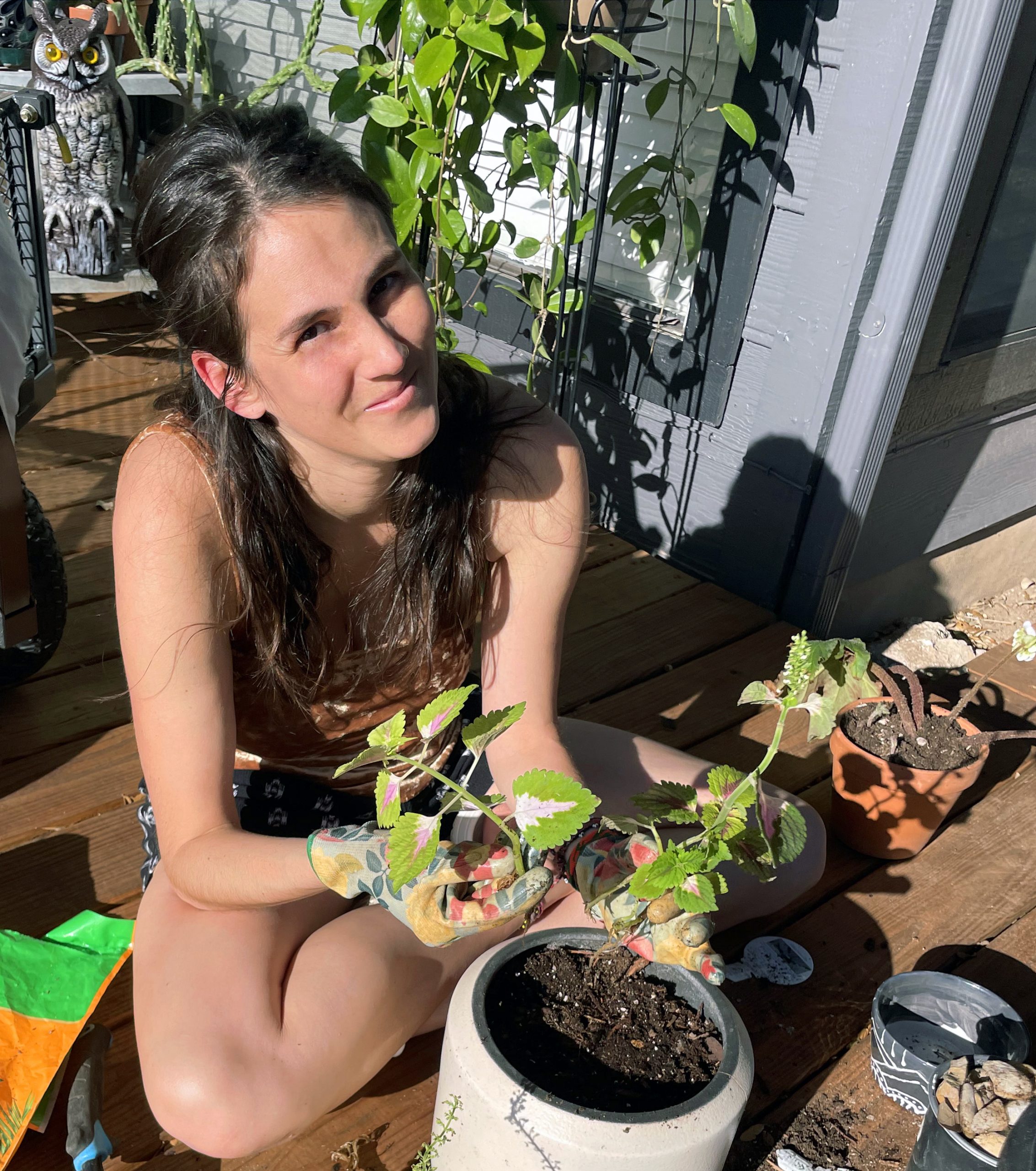 Steph Davlantes is sitting on a sunny porch wearing floral gardening gloves. She is surrounded by pots of soil and is holding two coleus plant babies.